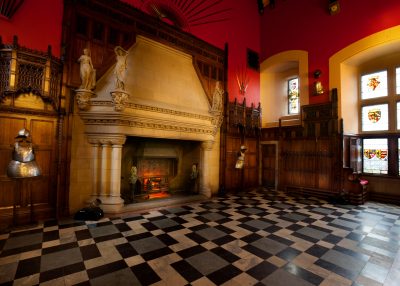 Image of a medieval castle with red walls, a mosaic floor, lit fireplace and suit of armour in the corner.
