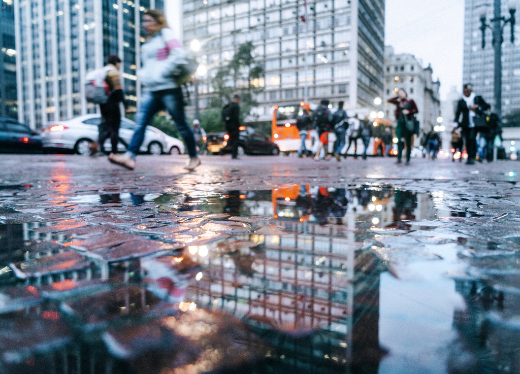 City scene of people walking through a rainy city, reflected in a puddle on an overcast day.